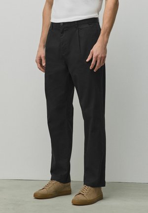 Man wearing black straight-leg trousers, beige lace-up sneakers, and a white t-shirt, standing against a light gray wall.