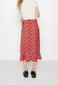 Jupe fleurie rouge avec un ourlet à volants, fabriquée en tissu léger. Équipée d'un détail de fermeture à nouer à la taille et de petits motifs floraux sur l'ensemble.