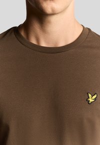 T-shirt di cotone marrone con scollo rotondo. Presenta un piccolo logo di un uccellino giallo ricamato sul lato sinistro del petto. Texture morbida e forma aderente.