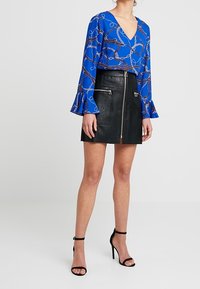 Modrá blůza s řetízkovým vzorem, výstřihem do V a zvonovými rukávy, spárovaná s černou koženou sukní se zipy vpředu a slim fit. Černé sandály na podpatku.