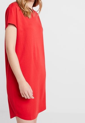 Frau mit heller Haut und blondem Haar, die ein lockeres rotes Kleid mit kurzen Ärmeln trägt und vor einem hellen Hintergrund steht.