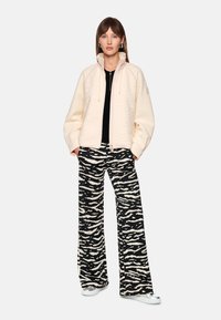 Light beige zip-up jacket with textured surface; paired with black and white striped wide-leg pants; wearing silver sneakers.