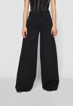Femme portant un jean noir taille haute à jambes larges et un haut noir sans manches avec des rayures blanches verticales sur un fond uni.