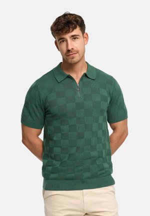 Mężczyzna o kręconych włosach, ubrany w zieloną krótko rękawową dzianinową koszulę w kratkę z kołnierzem na zamek, stojący z rękami za plecami.