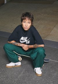 Černé oversized tričko s bílým logem Nike, zelené volné kalhoty, bílé tenisky s béžovou podrážkou, sedící na skateboardu. Betonové pozadí.