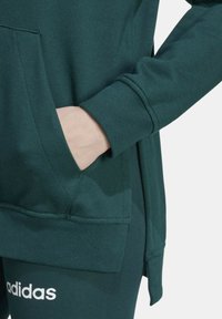 Persona che indossa un hoodie e pantaloni della tuta Adidas verde scuro, con la mano nella tasca dell'hoodie, su uno sfondo neutro.