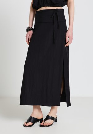 Donna che indossa una gonna nera fino alla caviglia con una spacco laterale, sandali neri con tacco aperto sulla punta e un top corto nero, in piedi davanti a uno sfondo neutro.