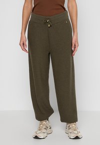 Olivengrüne, weit geschnittene Sweatpants mit einem strukturierten Muster. Sie haben einen elastischen Bund und einen Kordelzug und sind mit beigen Sneakern kombiniert.