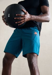 Turquoise athletic shorts with a subtle texture, featuring an orange logo. The individual holds a black weighted medicine ball.
