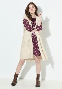 Beige Strickjacke über einem bordeauxfarbenen Blumenkleid, mit langen Ärmeln und plissierter Textur, kombiniert mit braunen Knöchelstiefeln.