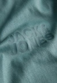 Ο τιρκουάζ ύφασμα διαθέτει μια υφή με ελαφρώς ανάγλυφο, ματ λογότυπο που γράφει "JACK & JONES" με έντονη γραμματοσειρά.