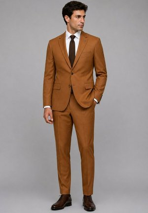 Homme debout portant un costume marron ajusté avec une chemise blanche, une cravate marron foncé et des chaussures habillées marron foncé sur un fond gris uni.