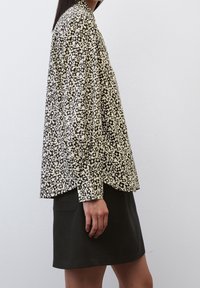 Camicia fluida a maniche lunghe color crema con motivo floreale nero, abbinata a una gonna verde scuro. Presenta un orlo arrotondato e polsini abbottonati.