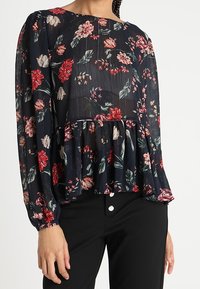 Blouse florale à manches longues, fond noir, fleurs rouges et roses, design plissé, texture douce, assortie à un pantalon noir avec des boutons blancs.