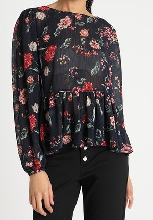 Blusa floreale a maniche lunghe, sfondo nero, fiori rossi e rosa, design plissettato, texture morbida, abbinata a pantaloni neri con bottoni bianchi.