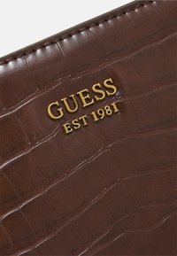 Brunt faux krokodillepræget materiale med en blank finish, der har det gyldne "GUESS EST 1981" logo præsenteret tydeligt.