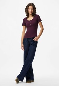 Jauna moteris su garbanotais plaukais, vilkinti purpurinę trumpą rankovę palaidinę, tamsias plačiąsias kelnes ir alyvuogių spalvos sportinius batelius, stovinti su sukryžiuotomis kojomis.