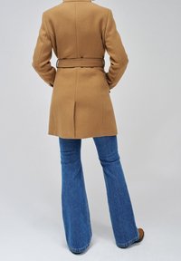 Cappotto di lana marrone chiaro con cintura, vestibilità sagomata e spacco singolo sul retro, abbinato a jeans denim blu svasati e calzature marroni.