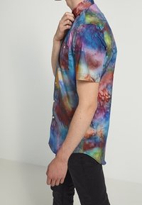 Košile s krátkým rukávem na knoflíky v duhovém vícebarevném batikovém vzoru. Má špičatý límec a náprsní kapsu na levé straně. Vyrobena z lehké látky.