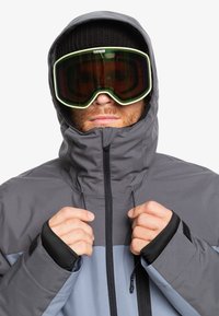 Grå och ljusblå vattentät jacka med hög krage, svarta ärmslut och justerbar huva, bärs tillsammans med stora gröntönta goggles.