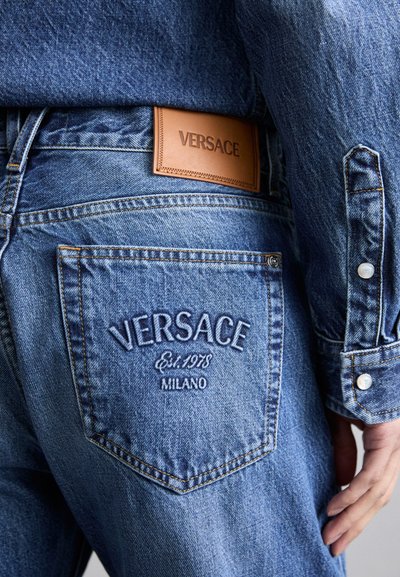 Mėlynos džinsinės kelnės turi odinį etikėtę su įspaudu "VERSACE" ant liemens ir siuvinėtų logotipą ant užpakalinės kišenės.