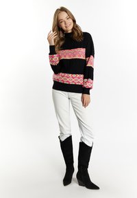 Pulover tricotat cu bază neagră, modele geometrice roz și galbene, guler înalt, asortat cu pantaloni albi și cizme din suede negre cu toc înclinat.