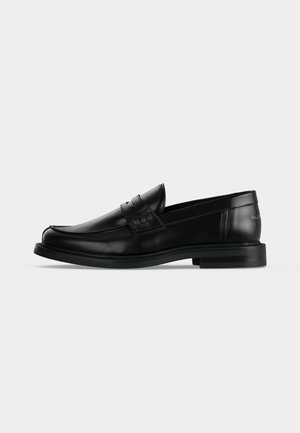 Mocasín negro de cuero con tacón bajo, una tira en la parte superior y detalles de costura sutiles sobre un fondo blanco liso.