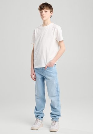 Camisa de algodón blanca, jeans de mezclilla azul claro y zapatillas blancas. Los jeans tienen un corte holgado con un diseño relajado y un detalle mínimo.