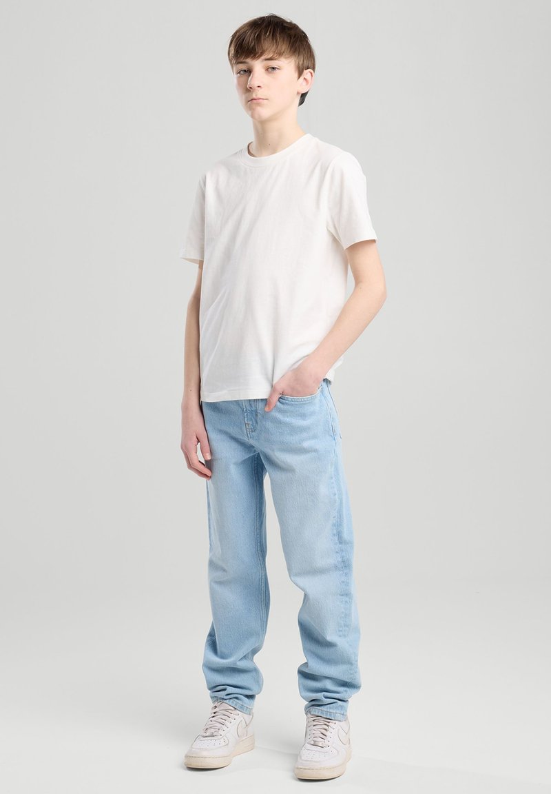 Valkoinen puuvillapaita, vaaleansiniset farkut ja valkoiset lenkkitossut. Farkut ovat väljät ja rentoa muotoilua sisältävät, ja niissä on minimaalinen yksityiskohtaisuus.