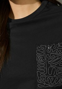 Sort t-shirt med en tekstureret lomme med sølvbroderede bølgede linjer. Glat stof med rund halsudskæring og korte ærmer.