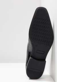 Semelle de chaussure formelle noire avec un grip texturé, détail de talon rectangulaire et marquage de taille "42", positionnée verticalement contre des surfaces blanches.