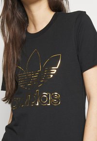 Svart bomulls t-shirt med en glänsande golden Adidas-logotyp, med kort ärm och rundad halsringning. Slät textur med en figurnära silhuett.