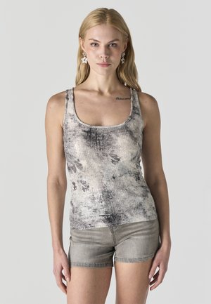 Mujer rubia con una camiseta sin mangas teñida estilo tie-dye en gris y negro, shorts de mezclilla grises y pendientes en forma de estrella, posando frente a un fondo gris claro.