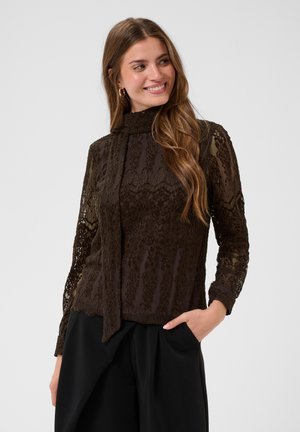 Jeune femme aux longs cheveux portant un chemisier en dentelle marron foncé à col haut et un pantalon noir, souriante et regardant sur le côté.