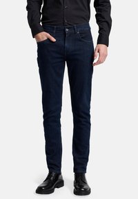 Mörkblå denimjeans med slim fit, med framfickor, knäppning och subtila sömnadsdetaljer. Bärs med svarta skor.