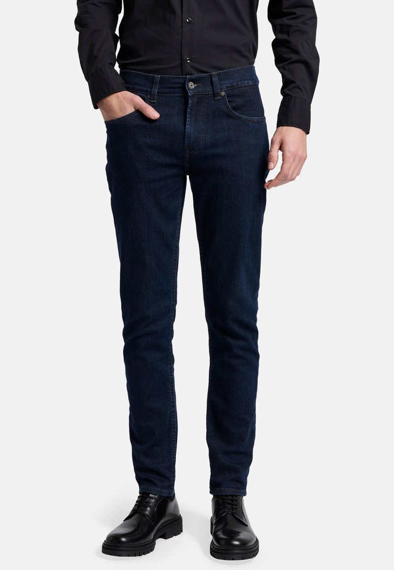 Mörkblå denimjeans med slim fit, med framfickor, knäppning och subtila sömnadsdetaljer. Bärs med svarta skor.