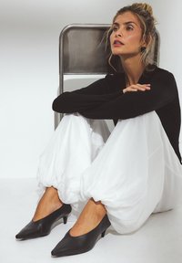Zapatos de tacón en punta de cuero negro con un tacón kitten bajo, usados con pantalones blancos fluidos, creando un contraste de colores y texturas.