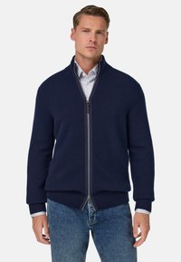 Marinblå zip-up tröja med ribbade ärmslut och nederkant, tillverkad av textil material; bärs över en blå och vit randig skjorta med låg krage.