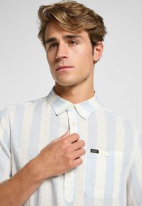 Camicia a righe in azzurro chiaro e bianco. Realizzata in tessuto leggero, presenta una tasca sul petto con un'etichetta del logo e bottoni a contrasto.