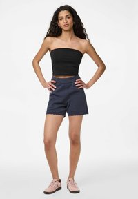 Haut court sans manches noir associé à un short texturé bleu marine avec des bords festonnés. Le mannequin porte des baskets rose clair. Arrière-plan simple.