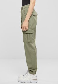 Khaki cargo kalhoty s elastickými manžetami, mnoha postranními kapsami a knoflíkovým zapínáním. Nosené s bílými teniskami, ukazující neformální styl.