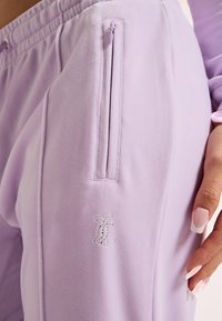 Približnji posnetek sivkasto vijoličnih trenirk z žepom na zadrgo na strani in monogramom iz kristalčkov, na katerem je vidna roka z francosko manikuro, naslonjena na bok.