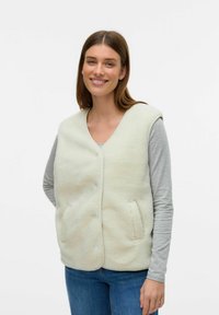 Gilet en fausse fourrure de couleur crème clair, avec un col en V, des boutons sur le devant et des poches latérales. Associé à un tee-shirt à manches longues gris et un jean bleu.