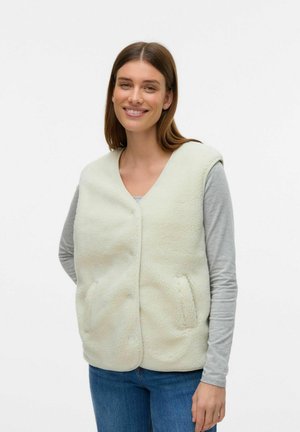 Faux-Fur-Weste in hellem Creme, mit V-Ausschnitt, vorderen Knöpfen und seitlichen Taschen. Kombiniert mit einem grauen Langarmshirt und blauen Jeans.