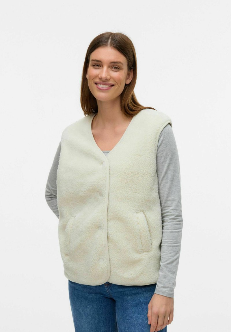 Gilet en fausse fourrure de couleur crème clair, avec un col en V, des boutons sur le devant et des poches latérales. Associé à un tee-shirt à manches longues gris et un jean bleu.