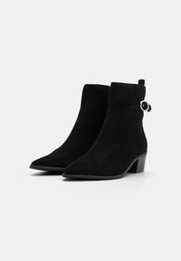 Botas de ante negras hasta el tobillo con puntera afilada, tacón bajo y un pequeño detalle de correa con una hebilla redonda de plata. Textura suave.