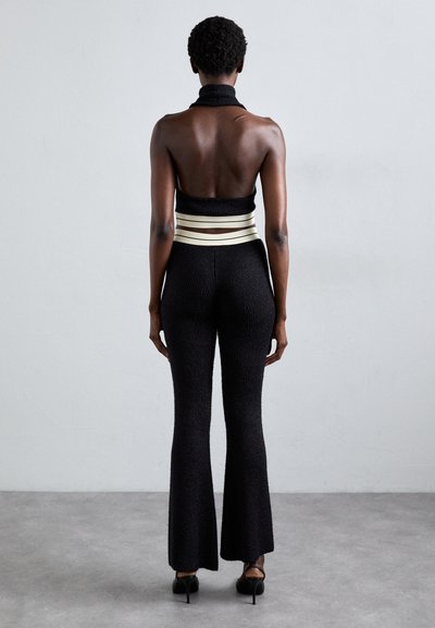 Woman standing facing away wearing black textured flared pants, black halter top with wide cream cutout bands, and black heels on gray floor.