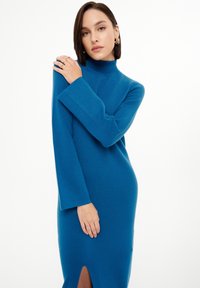 Langärmliges, Turtleneck-Kleid aus lebhaft blauer Strickware mit seitlichem Schlitz, glatter Textur und lockerer Passform; das Modell trägt es mit einer Hand auf ihrer Schulter.