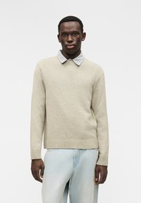 Μπεζ πλεκτό πουλόβερ με ριμπ τελείωμα και μανσέτες, φορεμένο πάνω από πουκάμισο με καρό γιακά. Το ύφασμα φαίνεται μαλακό και με υφή.