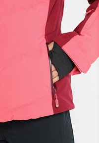 Main partiellement dans la poche zippée latérale d'une veste bicolore rose et bordeaux avec une manche intérieure noire visible.